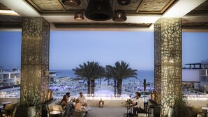 Een terras buiten met uitzicht op het strand en palmbomen, met tafels en stoelen, en een podium met muziekinstrumenten.