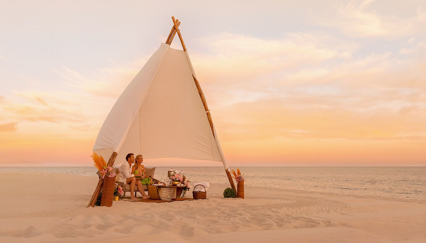 A couple sitting under a large white sail tent on a beach at sunset.