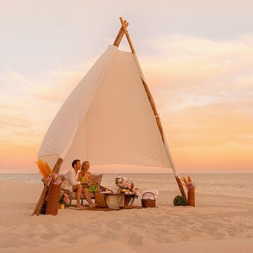 A couple sitting under a large white sail tent on a beach at sunset.