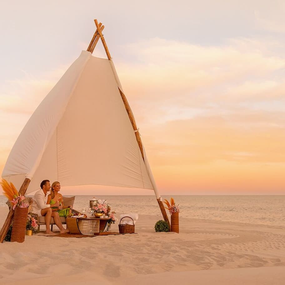 A couple sitting under a large white sail tent on a beach at sunset.