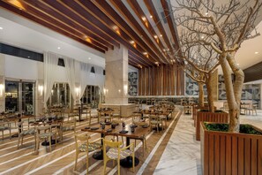A spacious dining area with wooden tables and chairs, a large tree, and a modern ceiling design.
