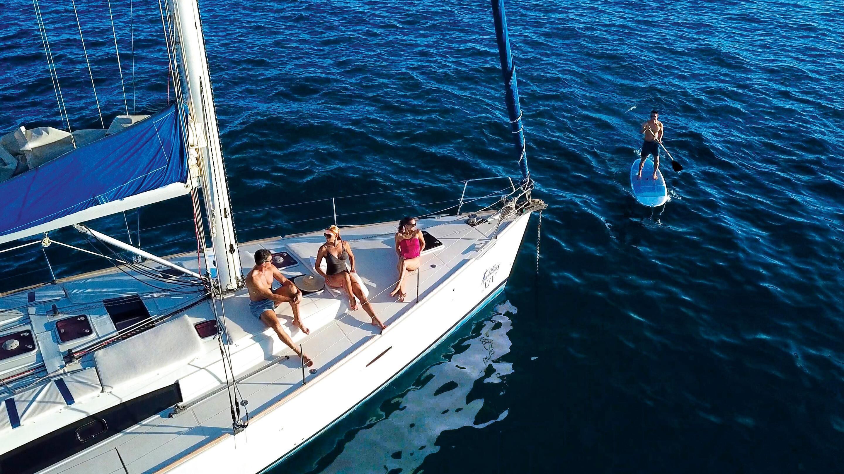 Un velero con tres personas en la cubierta, una persona remando en el agua y una vista despejada del océano.