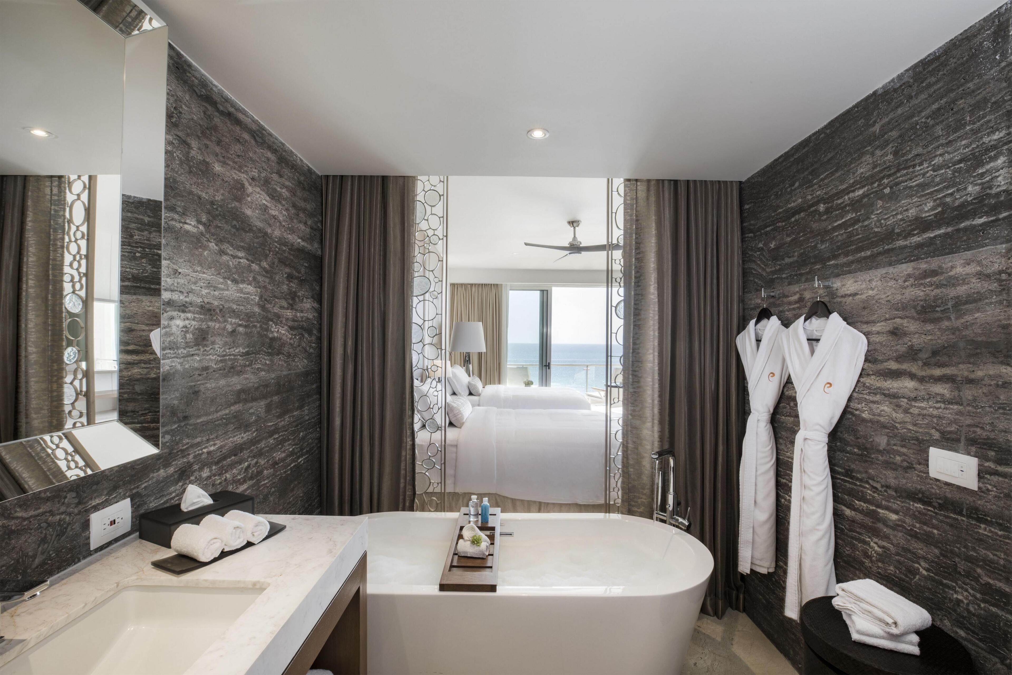 Een moderne badkamer met een groot bad, een wastafel met spiegel en uitzicht op een slaapkamer met een bed en een raam.
