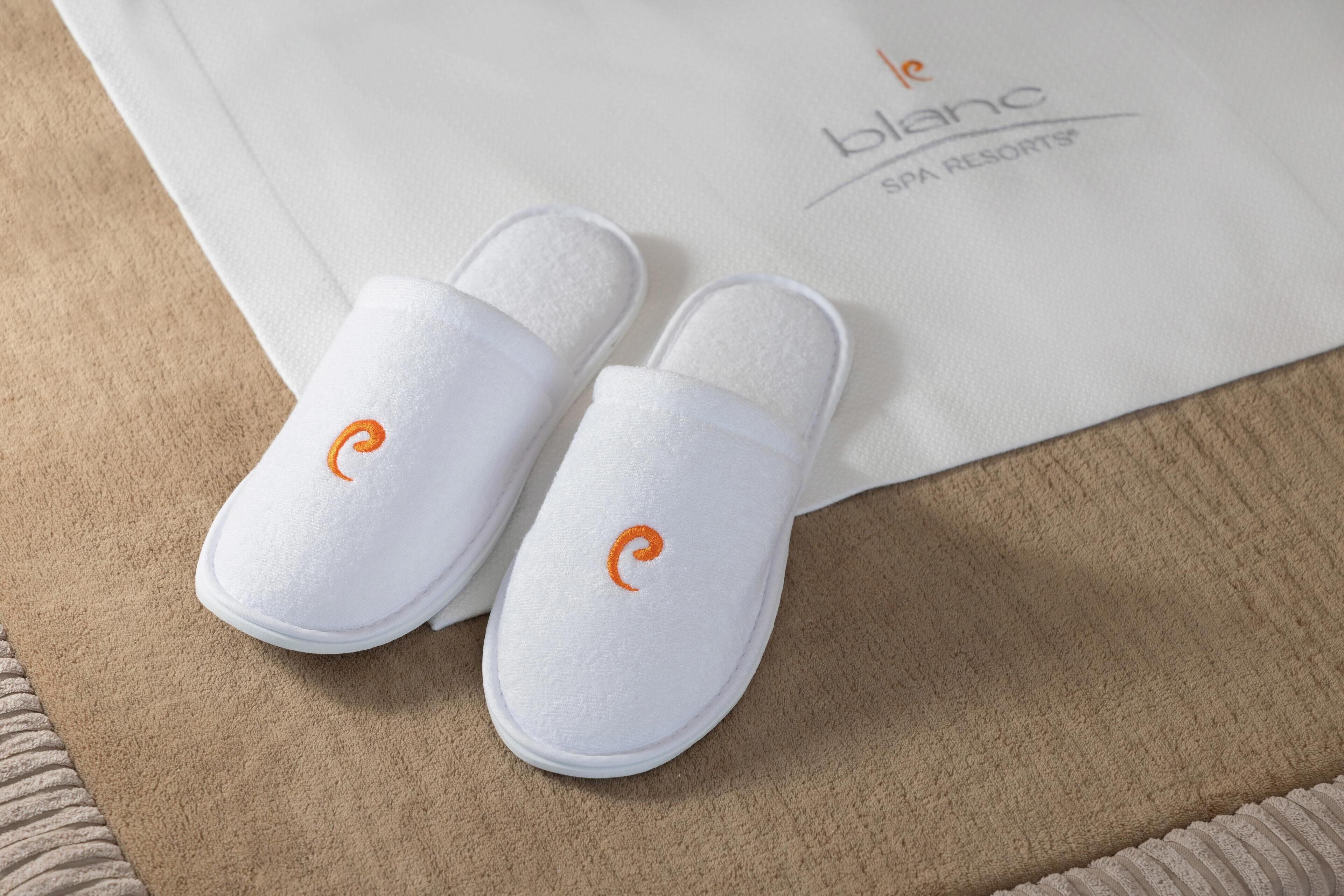 A pair of white slippers with an orange logo on a beige carpet, next to a white paper with text.