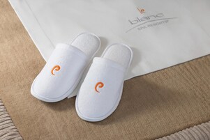 A pair of white slippers with an orange logo on a beige carpet, next to a white paper with text.