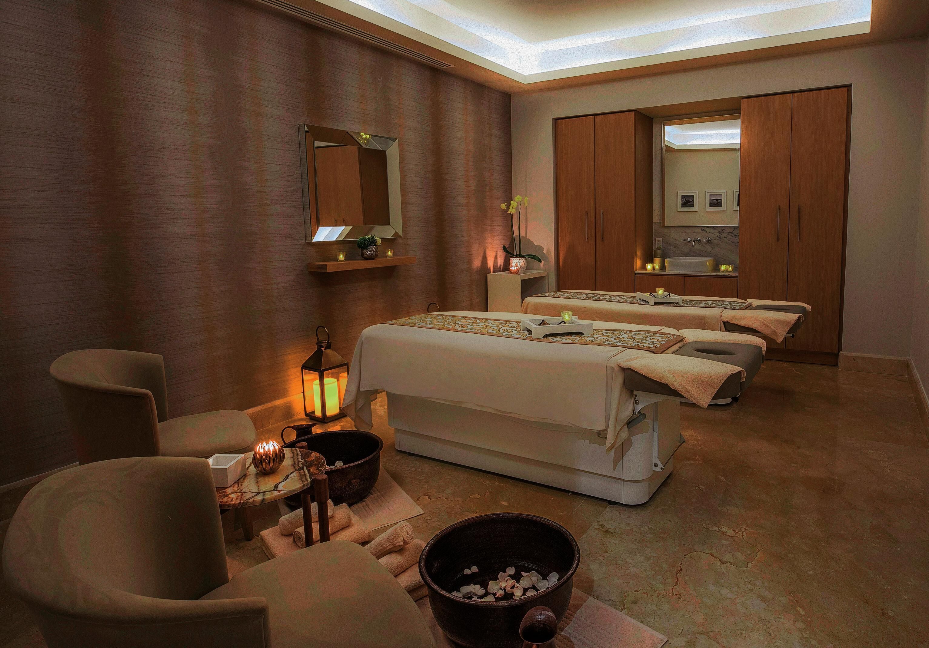 Een spa-ruimte met drie massagetafels, een zithoek met stoelen, een tafeltje met een kaars, en een grote schaal met stenen.