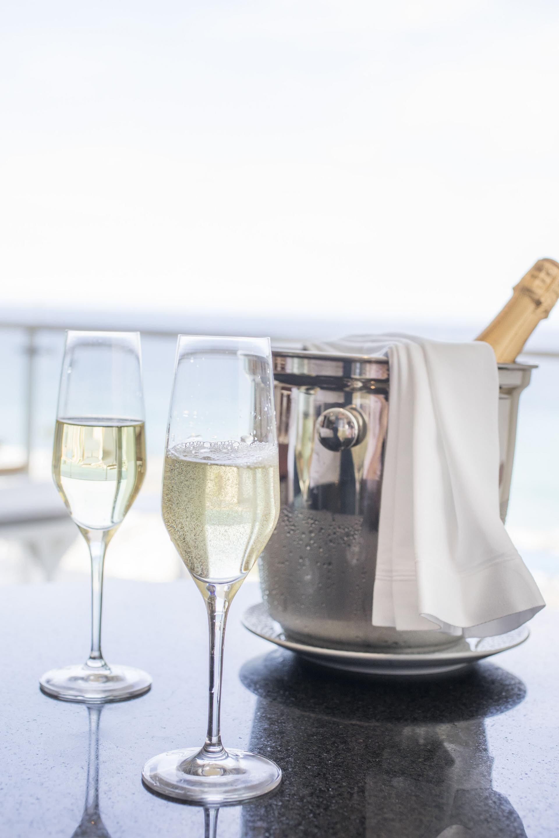 Two champagne flutes on a reflective surface with a champagne bottle and a metal container in the background.