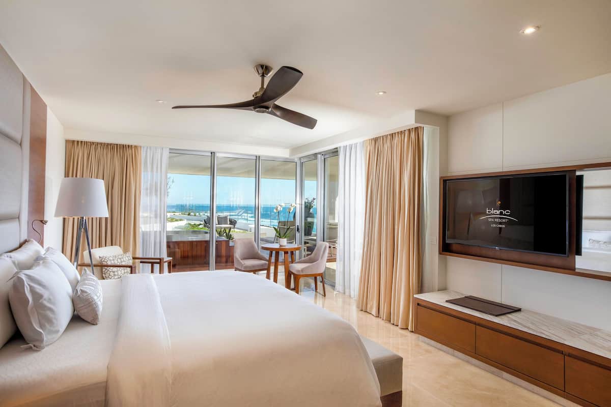 Une chambre d’hôtel équipée d’un lit, d’une télévision, d’un ventilateur de plafond, d’un balcon avec vue et d’un bureau avec des chaises.