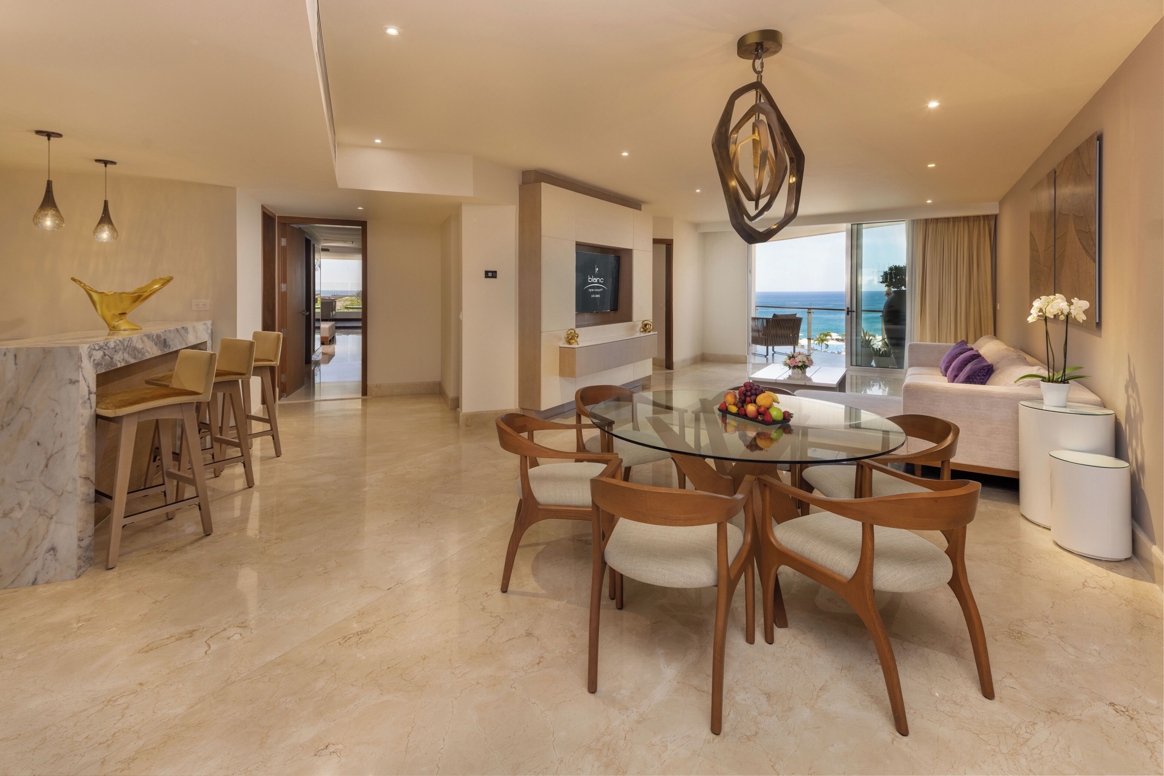 A modern living room with a glass dining table, wooden chairs, a marble fireplace, and a view of the sea.