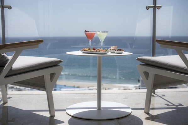 A balcony with two white chairs, a small round table with a plate of food and two drinks, and a view of the ocean.