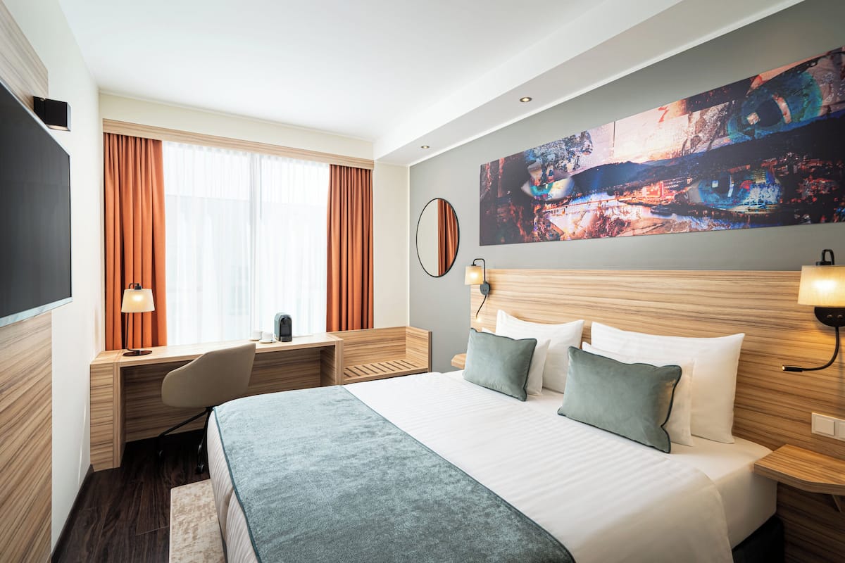 Hotellihuoneessa on suuri sänky, työpöytä ja tuoli. Seinällä on taulu, ikkunassa verhot ja seinällä myös televisio.