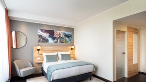 A modern hotel room with a large bed, a chair, a bedside table, a wall-mounted painting, and a door. - Leonardo Boutique Hotel Linz City Center (Linz)