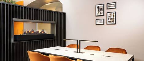 A modern conference room with a long table, orange chairs, a fireplace, and framed pictures on the wall.