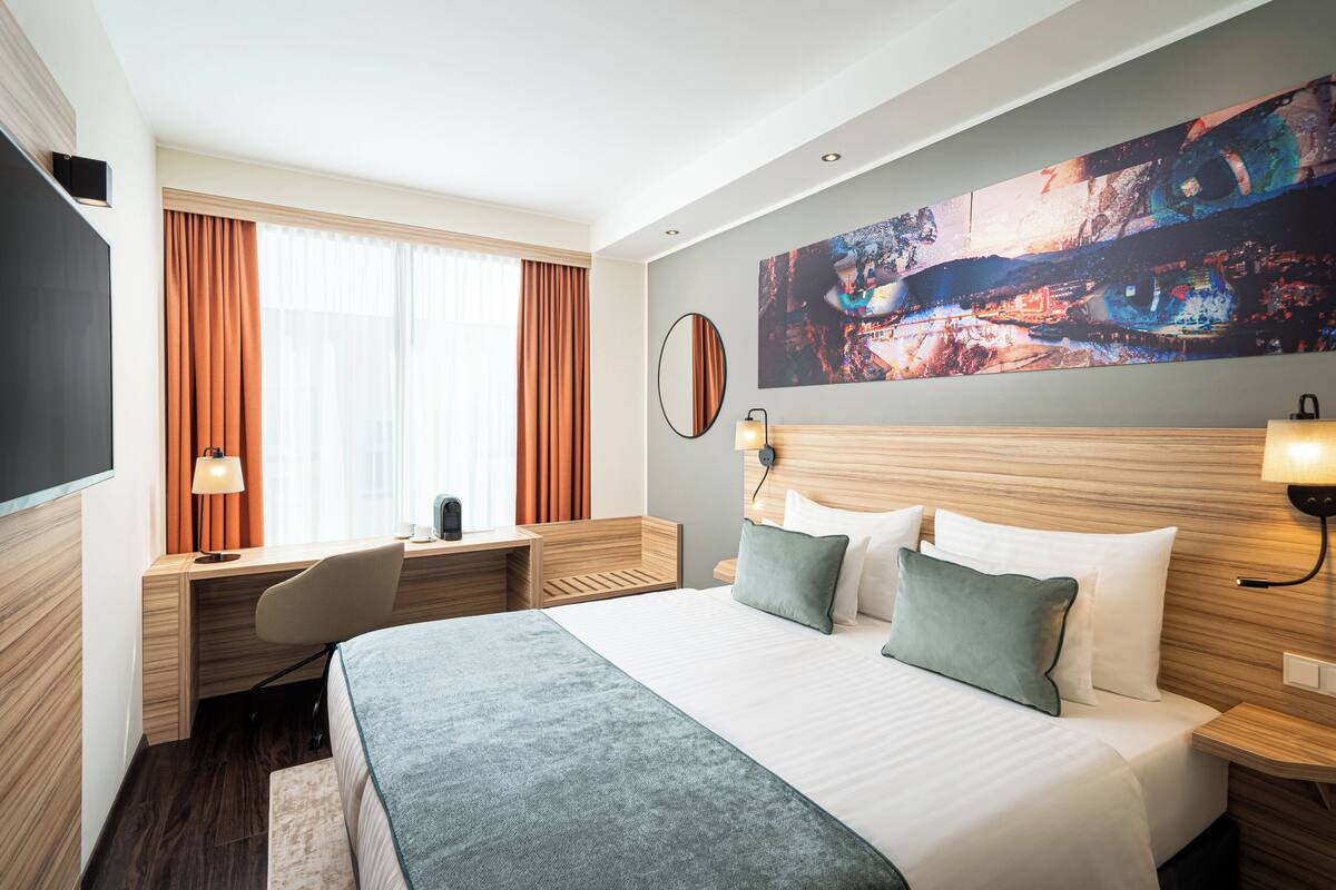 Una habitación de hotel con una cama grande, un escritorio y una silla. Hay un cuadro en la pared, una ventana con cortinas y un televisor también en la pared.