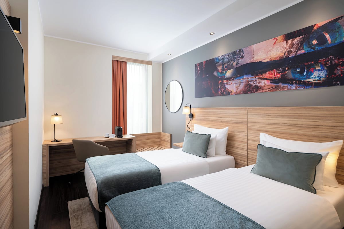Habitación de hotel con dos camas, un escritorio, una silla, un televisor, un cuadro y una ventana con cortinas.