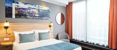 A modern hotel room with a large bed, a cityscape painting, a round mirror, and a view of the outdoors through sheer curtains.