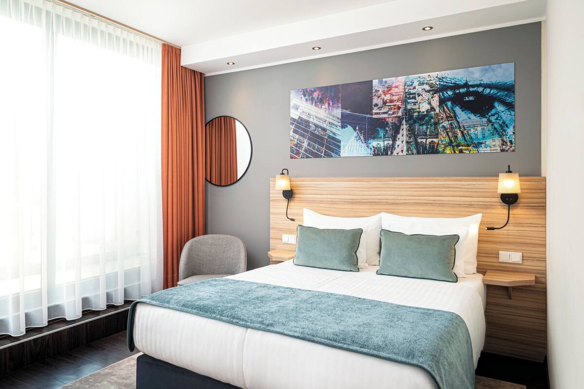 Una habitación de hotel moderna con una cama grande, un cabecero con luces empotradas, un espejo redondo y una pintura de un paisaje urbano en la pared.