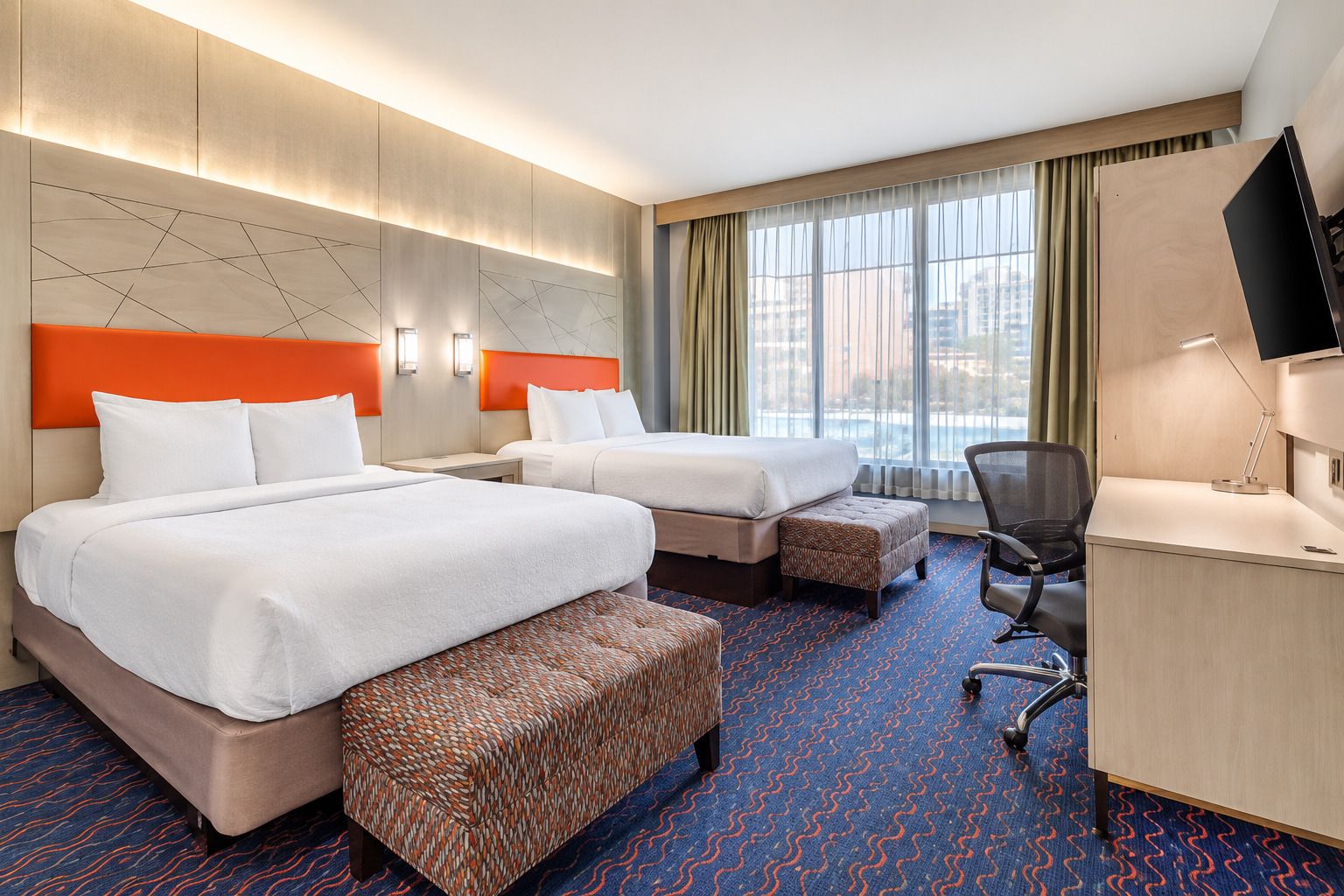 Une chambre d’hôtel avec deux lits, un bureau, une chaise et une grande fenêtre avec des rideaux.