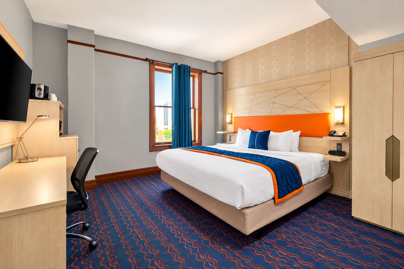 A hotel room with a bed, desk, chair, and a view of the outdoors.