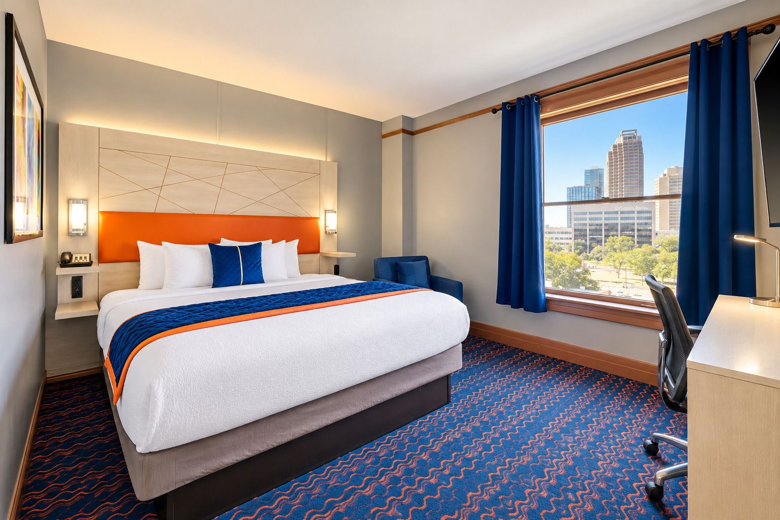 A hotel room with a large bed, a desk, a chair, a view of the city, and a modern headboard.