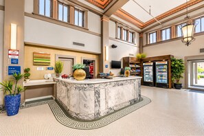 Un espace d’accueil d’hôtel avec un comptoir en marbre, une grande plante en pot, un réfrigérateur et un téléviseur.
