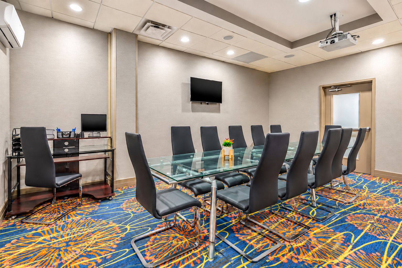 A conference room with a glass table, black chairs, a TV on the wall, and a carpeted floor.