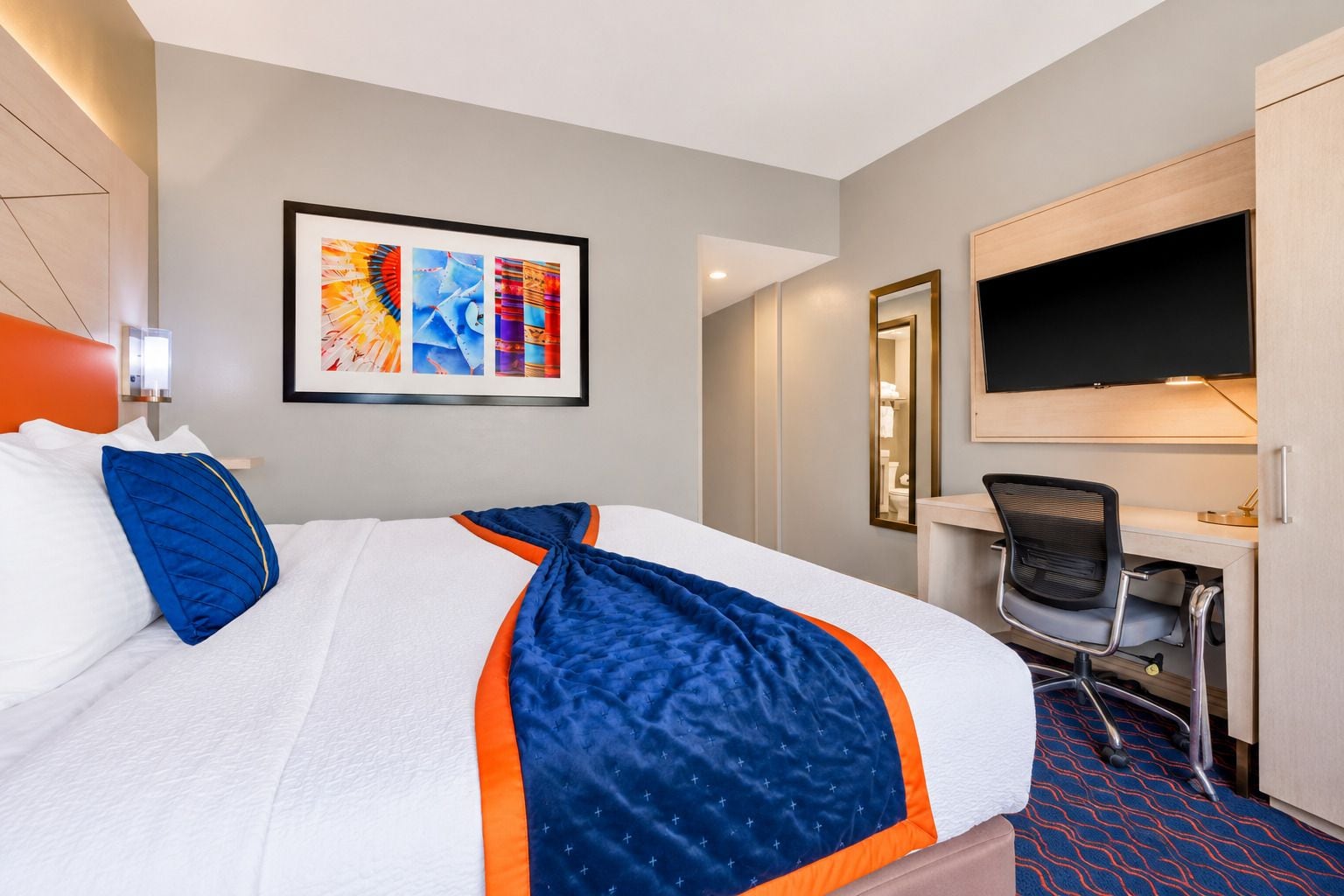 A hotel room with a bed, a desk, a chair, a TV, and a framed artwork on the wall.