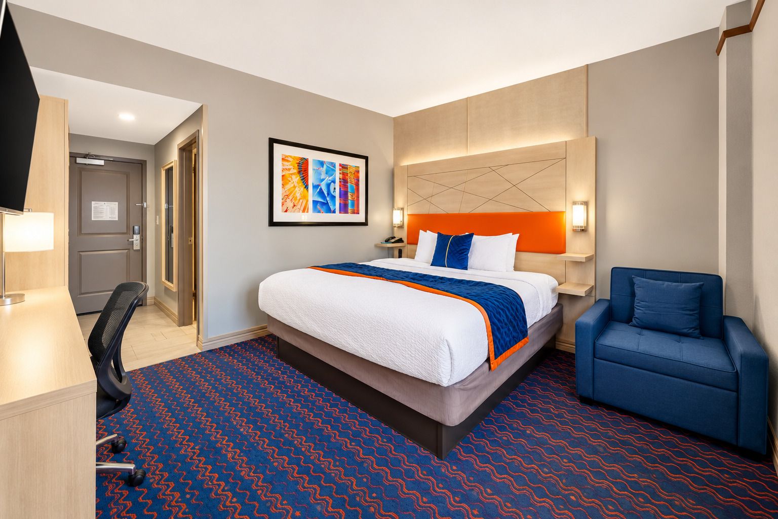 A hotel room with a bed, a desk, a chair, a wall-mounted TV, and a framed picture on the wall.