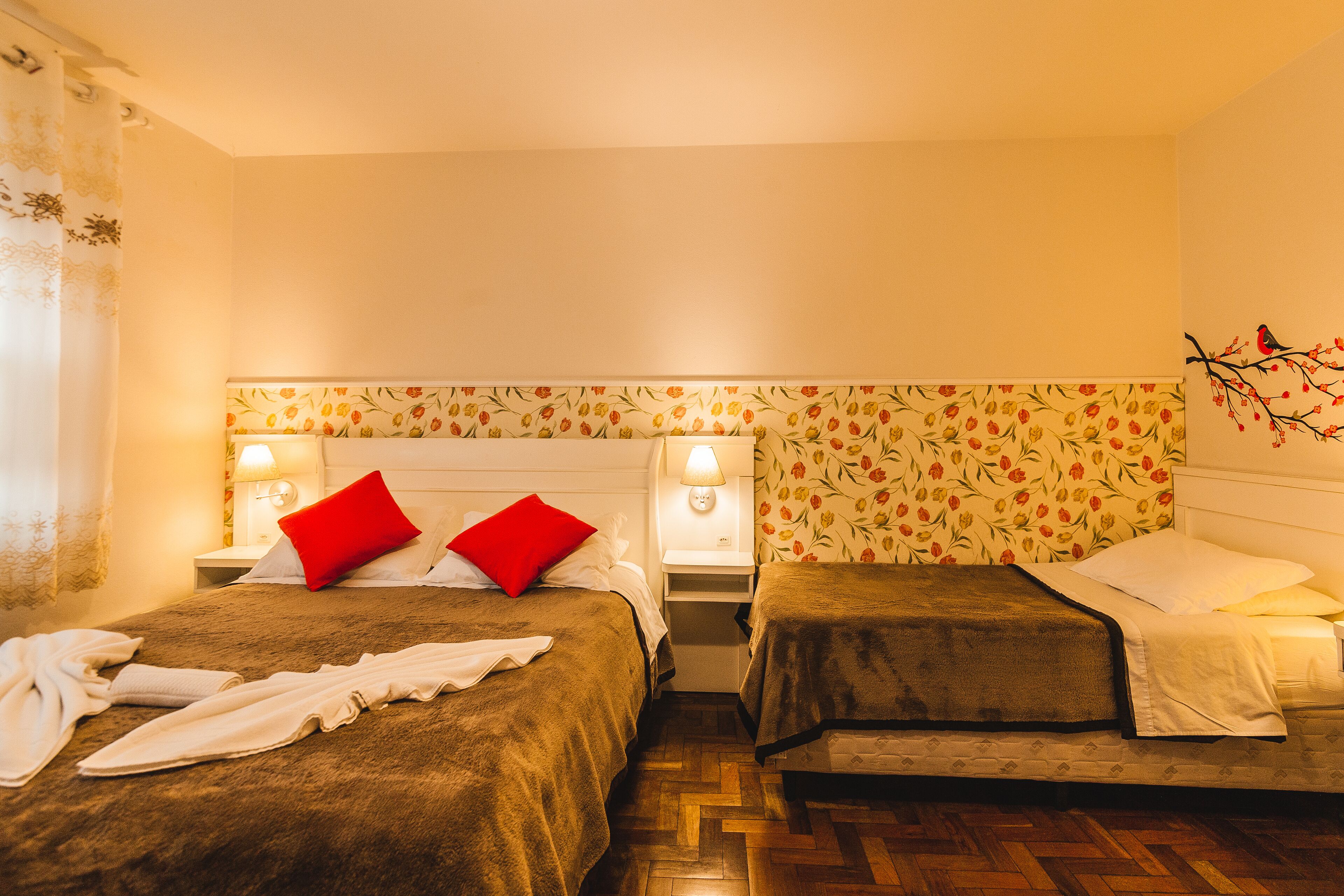 A hotel room with two beds, a wooden floor, floral wallpaper, and bedside lamps.