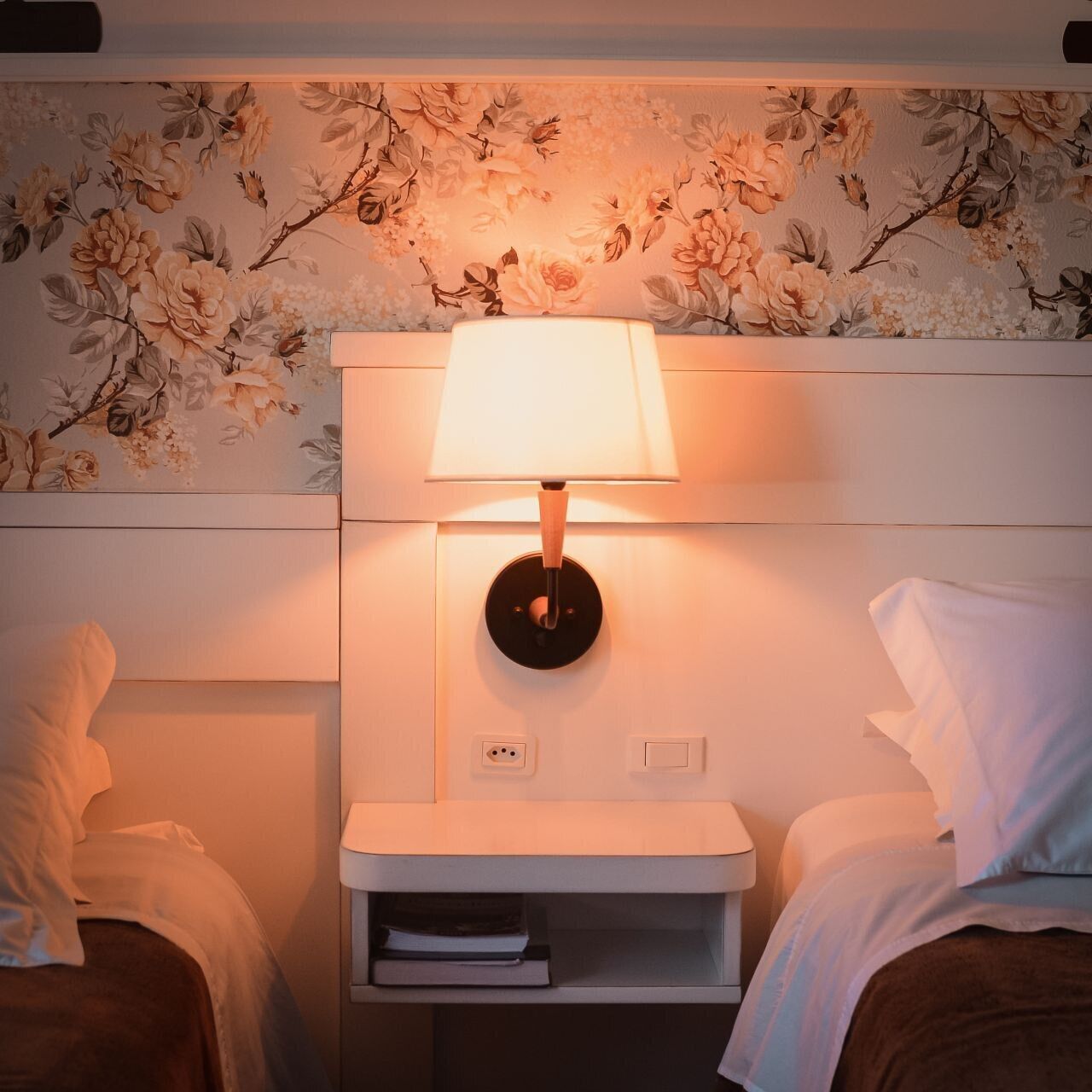 A hotel room with two beds, a nightstand with a lamp, and a floral wallpaper design.