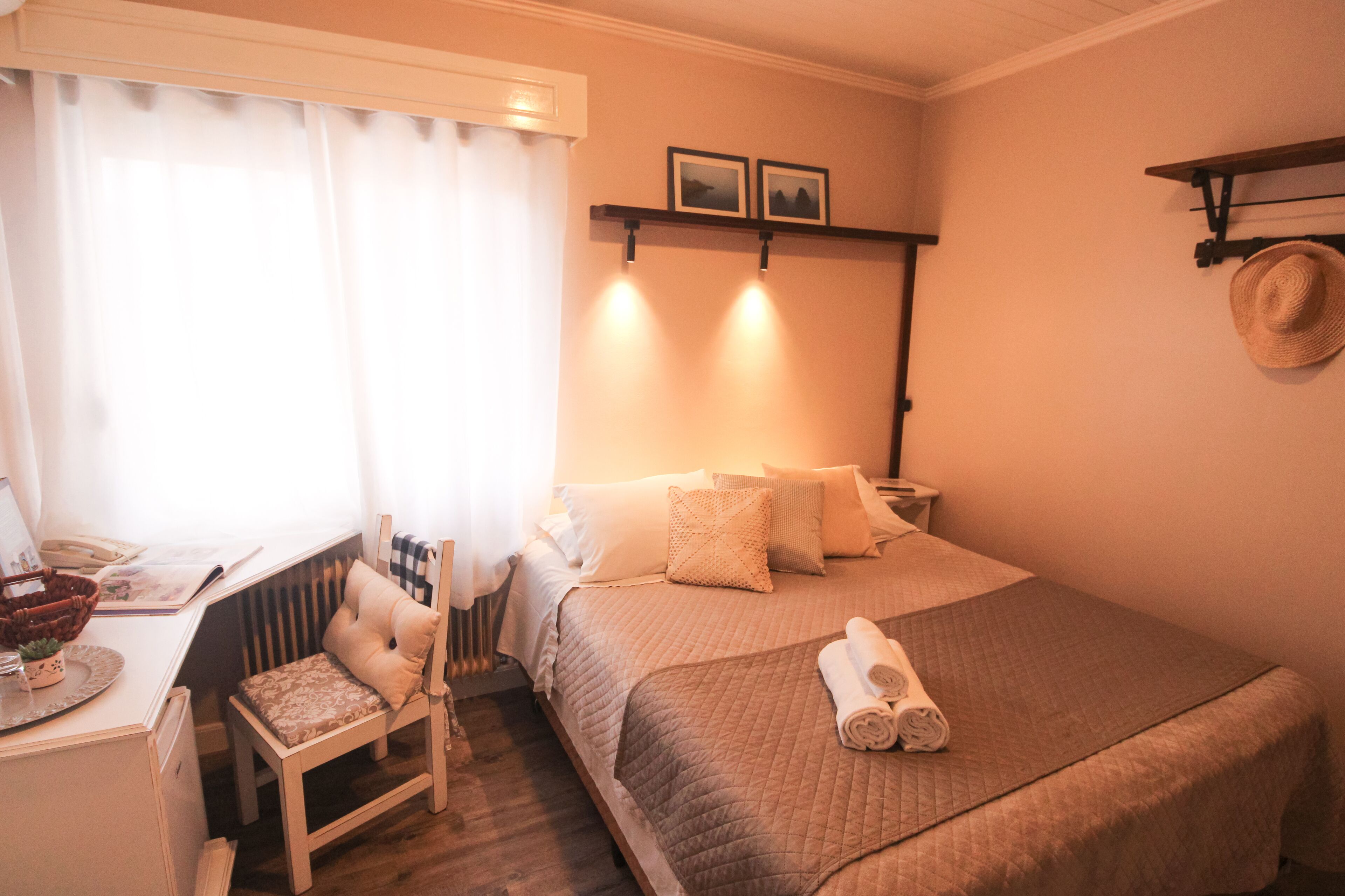 A neatly arranged bedroom with a bed, a chair, a desk, and a shelf with a hat.