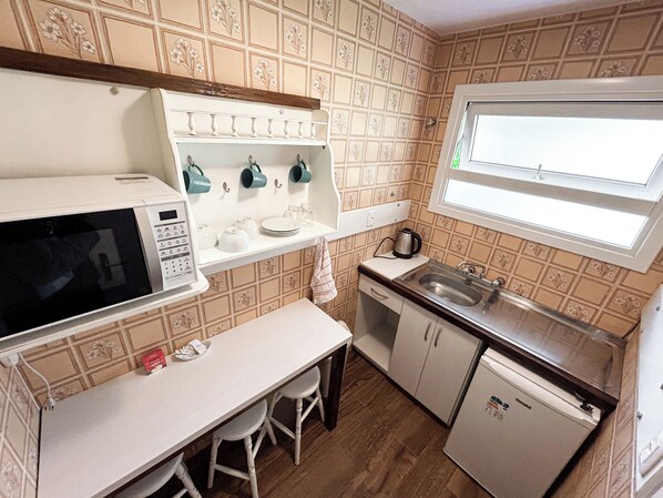 A compact kitchen with a microwave, sink, and small refrigerator. There are stools and a countertop with a window above the sink. - Pousada Betânia (Gramado)