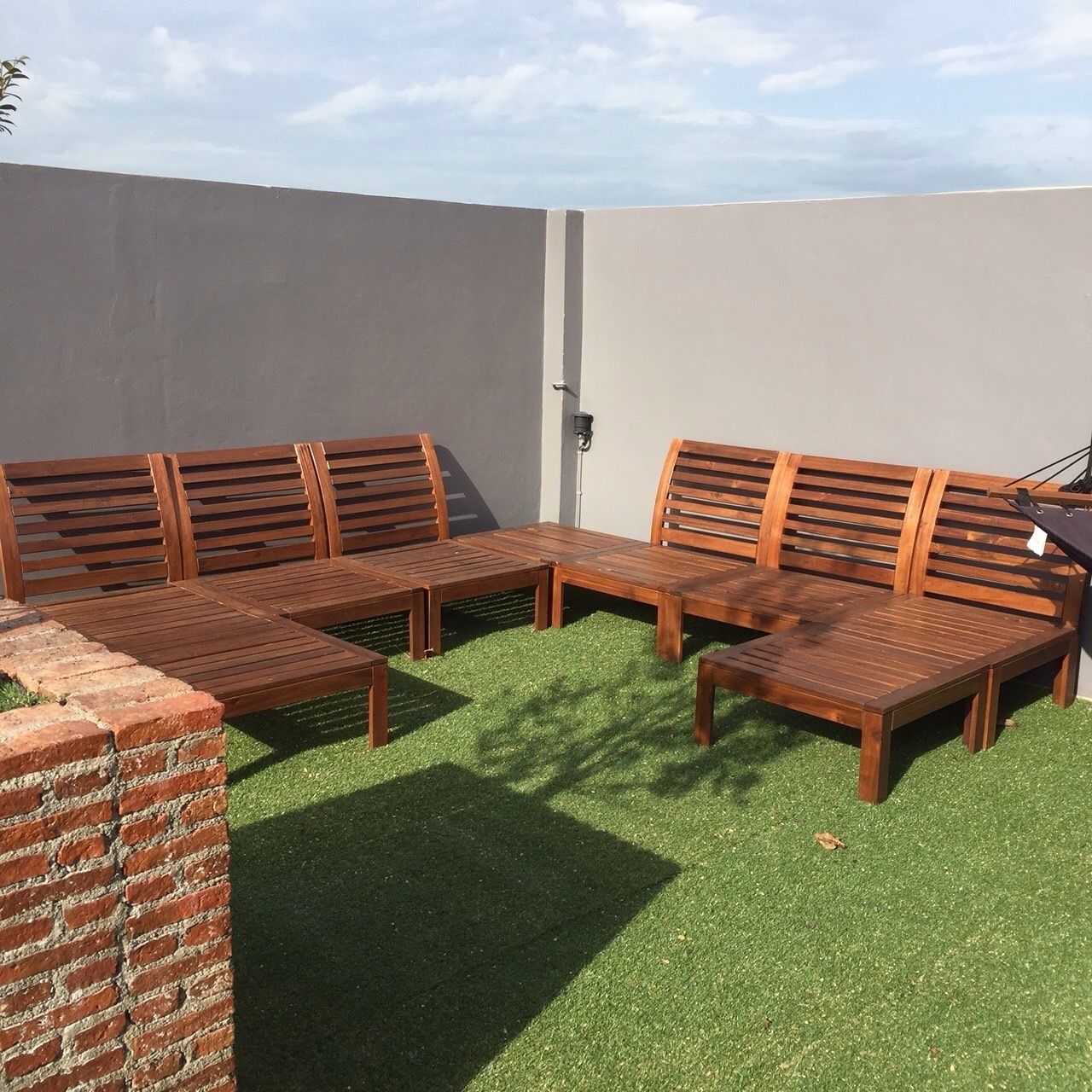 A wooden bench set on artificial grass with a brick wall and a plain white wall in the background.