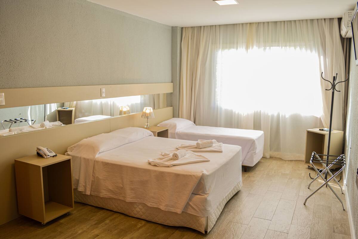 Une chambre d’hôtel avec deux lits, une table de chevet avec une lampe, un miroir, un téléphone et une fenêtre avec des rideaux.