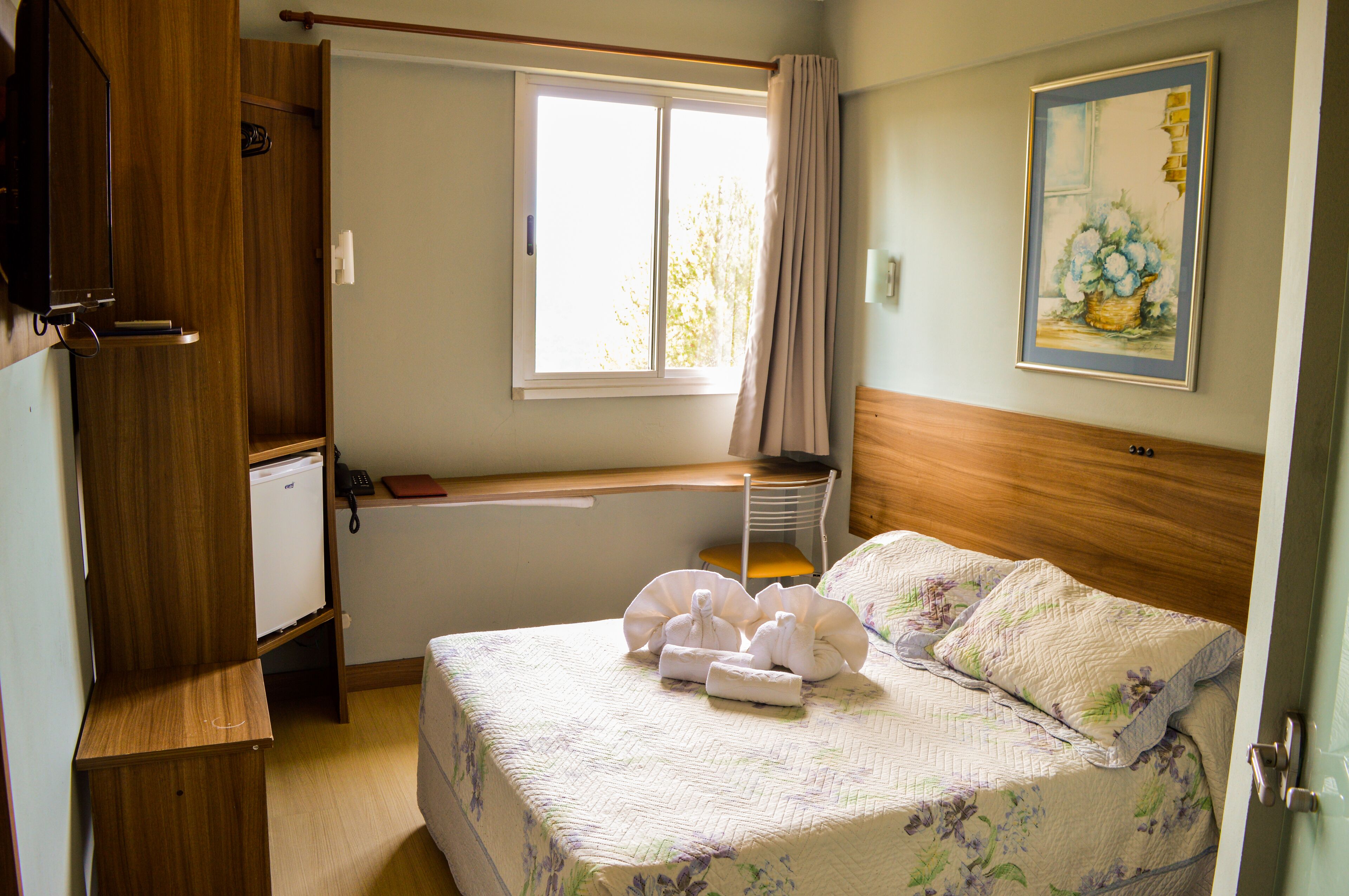 A bedroom with a bed, a window, a desk, and a small refrigerator.