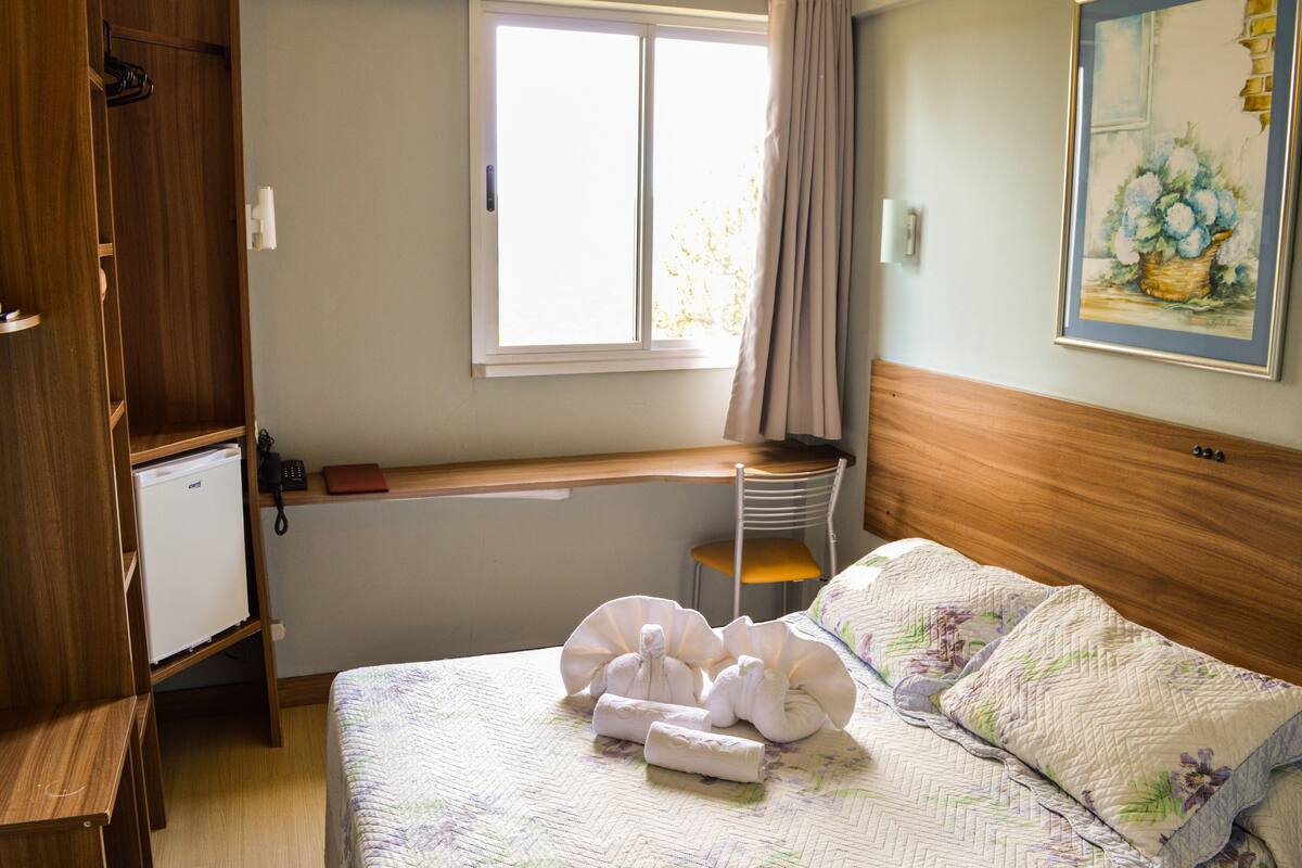 Une chambre d’hôtel avec un lit, un petit bureau, une chaise, une fenêtre avec des rideaux et un tableau au mur.