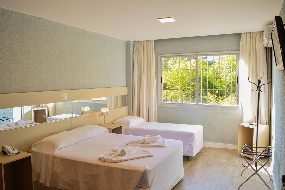 Une chambre d’hôtel avec deux lits, une grande fenêtre donnant sur un espace verdoyant, un miroir, une table de chevet avec une lampe et un téléphone.