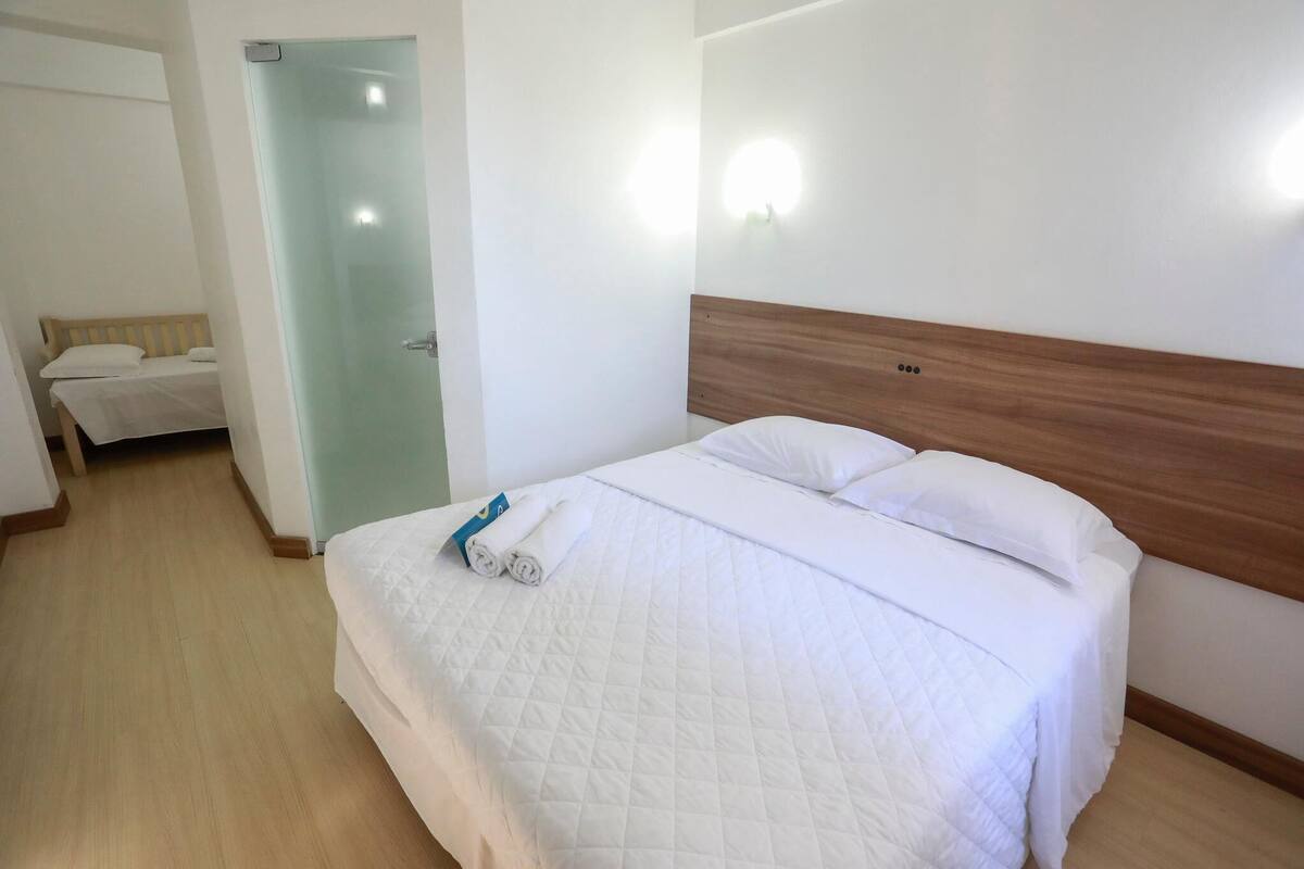 Une chambre d’hôtel avec un lit, une tête de lit en bois, une lampe fixée au mur et une porte donnant sur une autre pièce.