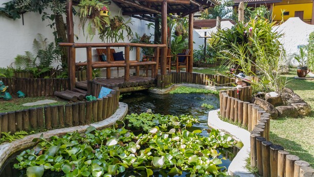 Eine Holnpergola mit Sitzgelegenheit, ein kleiner Teich mit Seerosen und umgebende Bepflanzung.
