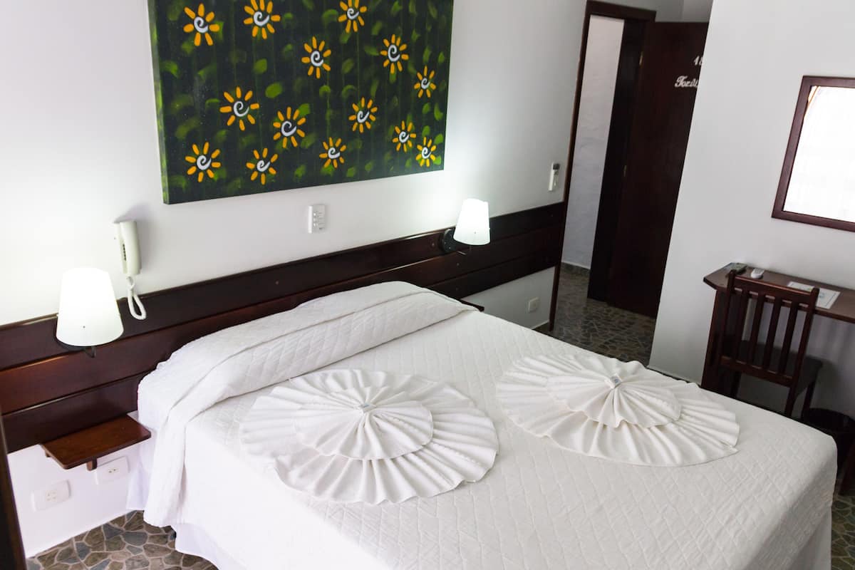 Habitación de hotel con cama, mesita de noche, lámpara, un cuadro en la pared y una puerta al fondo.