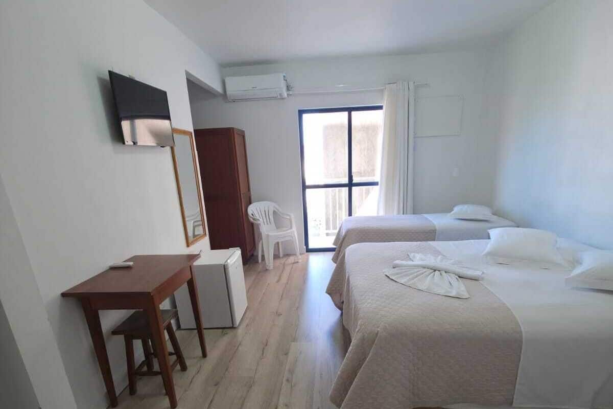 A hotel room with a desk, two beds, a chair, and a large window.