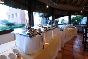 Montaje de bufé con manteles blancos y cubiertos de plata, un florero con flores y vistas al exterior con vegetación.