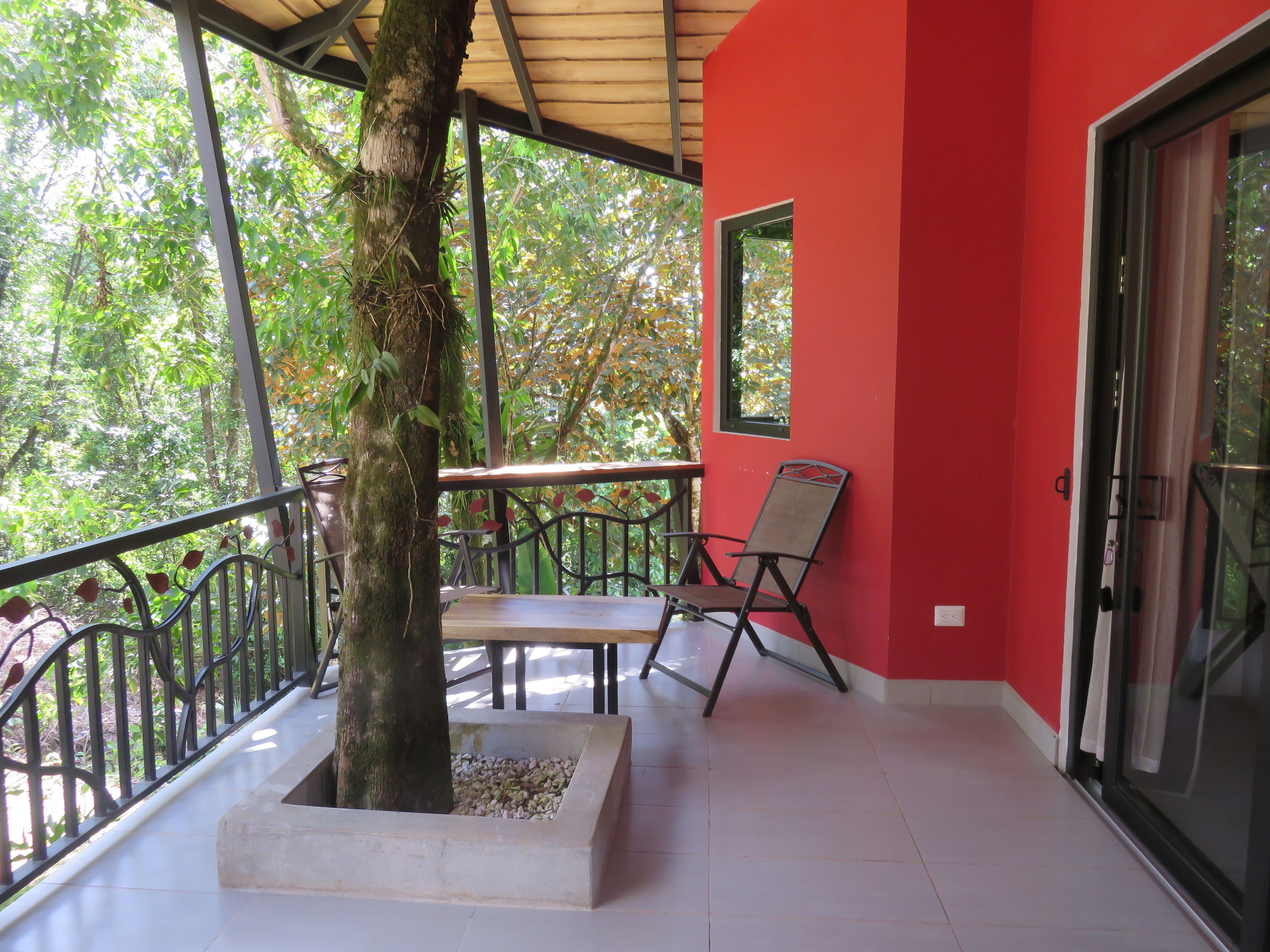 A covered balcony with a table and chairs, a tree planter, and a red wall.