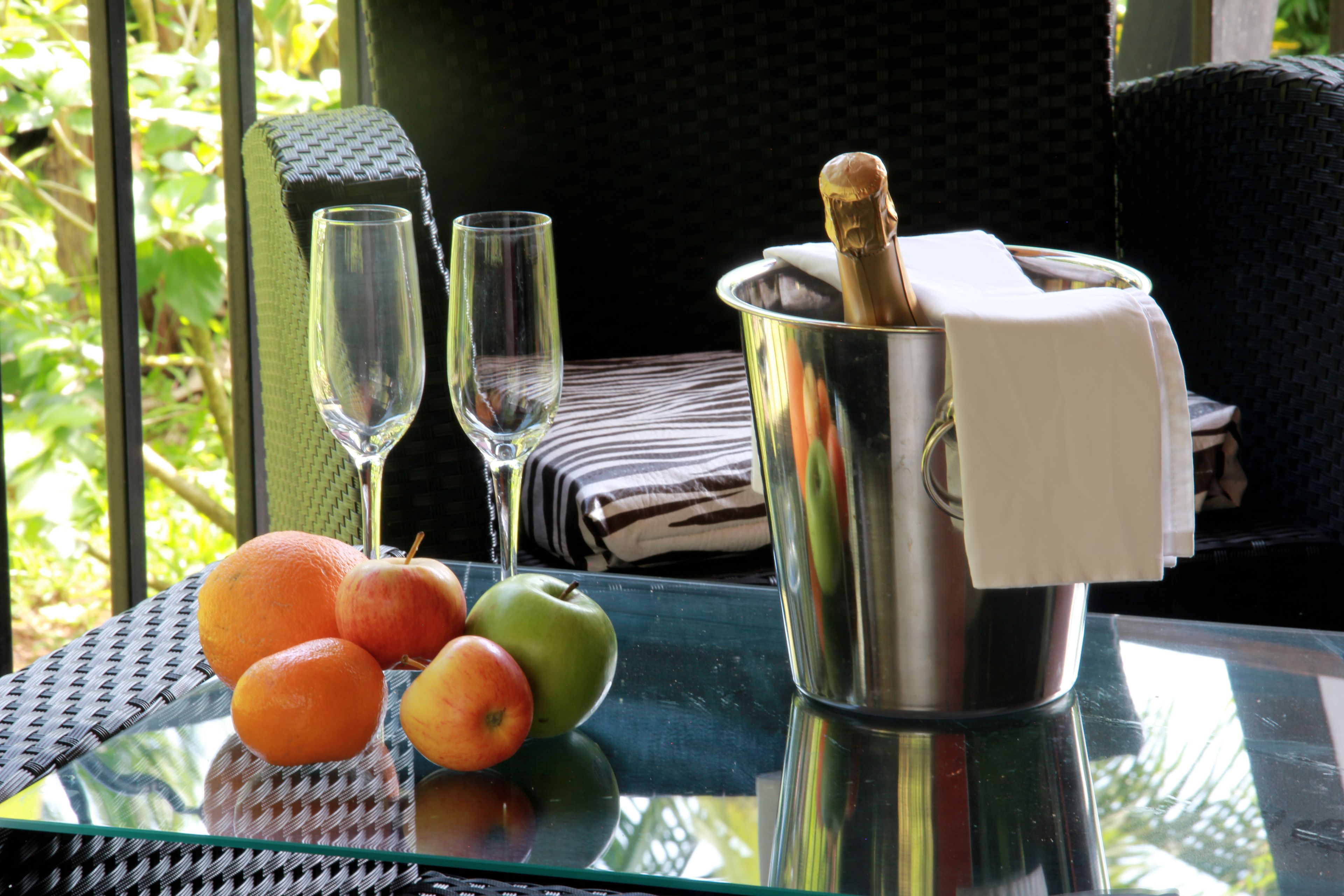 A glass table with a bottle of champagne, two glasses, and three fruits.