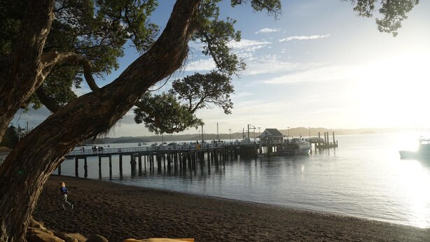 A beach with a pier extending into calm waters, a large tree in the foreground, and a person walking on the sand. - Kokomo Retreat (Russell)
