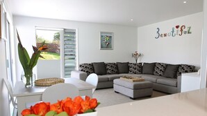 A modern living room with a grey sofa, a white coffee table, and a dining area with a white table and chairs. There is a large window with blinds, a painting on the wall, and a decorative word "Beautiful" above the sofa. - Kokomo Retreat (Russell)