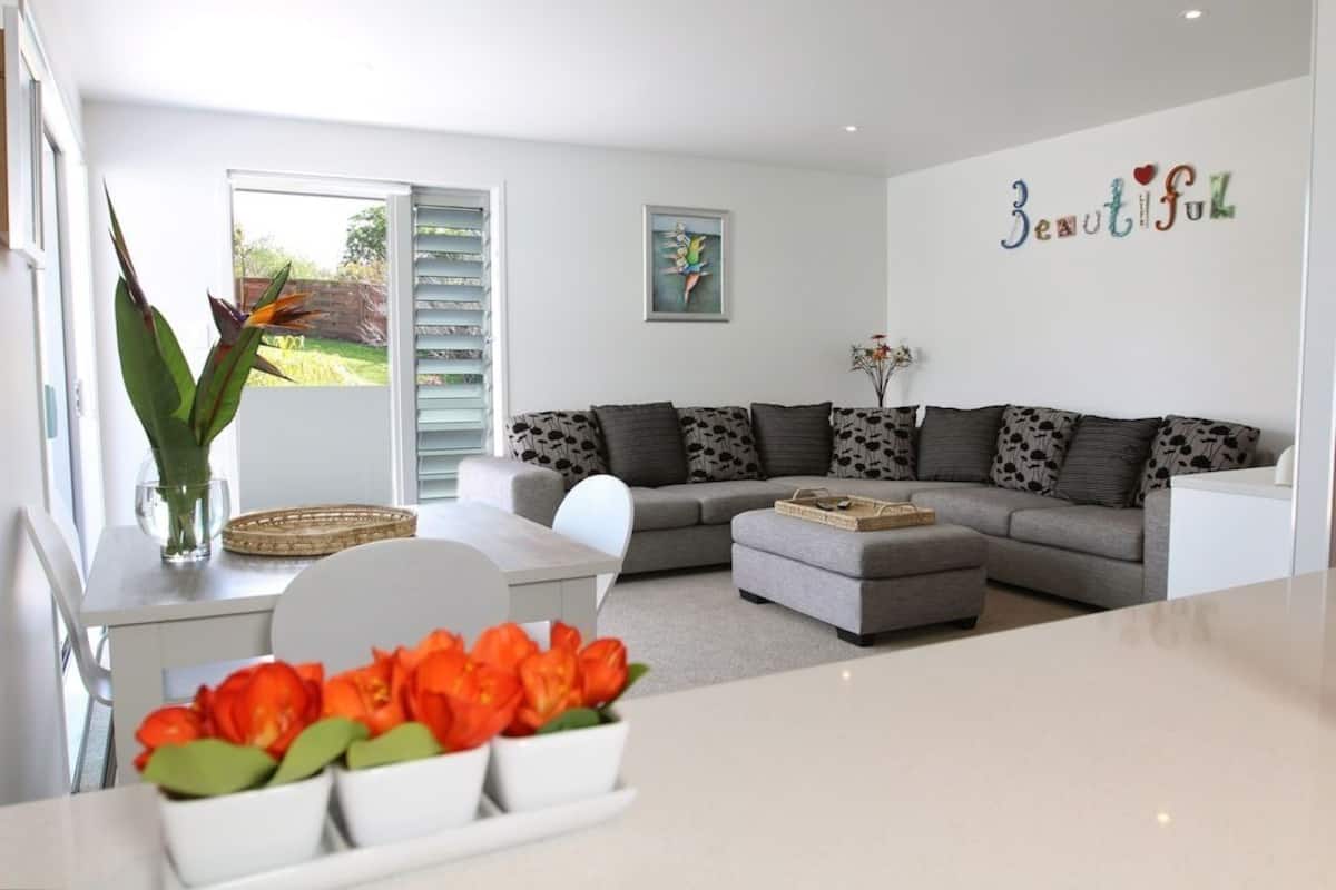 Un salon moderne avec un canapé gris, une table basse blanche et un coin repas comprenant une table et des chaises blanches. On y trouve une grande fenêtre avec des stores, un tableau au mur et le mot décoratif «
Beautiful
» au-dessus du canapé.