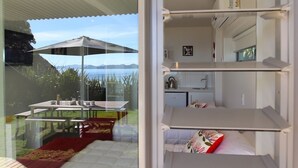 A modern, compact room with a glass wall offering a view of the outdoors. It includes a dining area with a table and chairs, a kitchenette with a sink and stove, and a bed with a red accent pillow. - Kokomo Retreat (Russell)