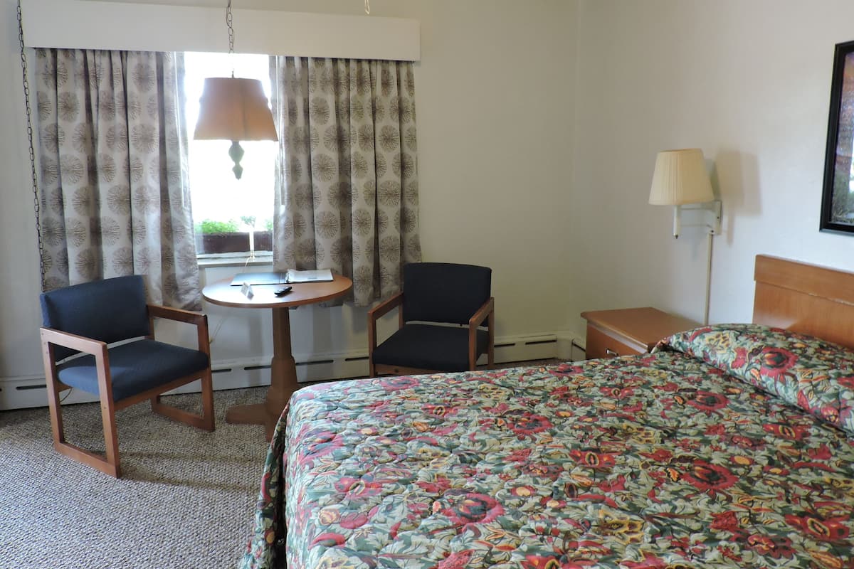 Una habitación de hotel con cama, dos sillas, una mesita, una lámpara y ventana con cortinas.