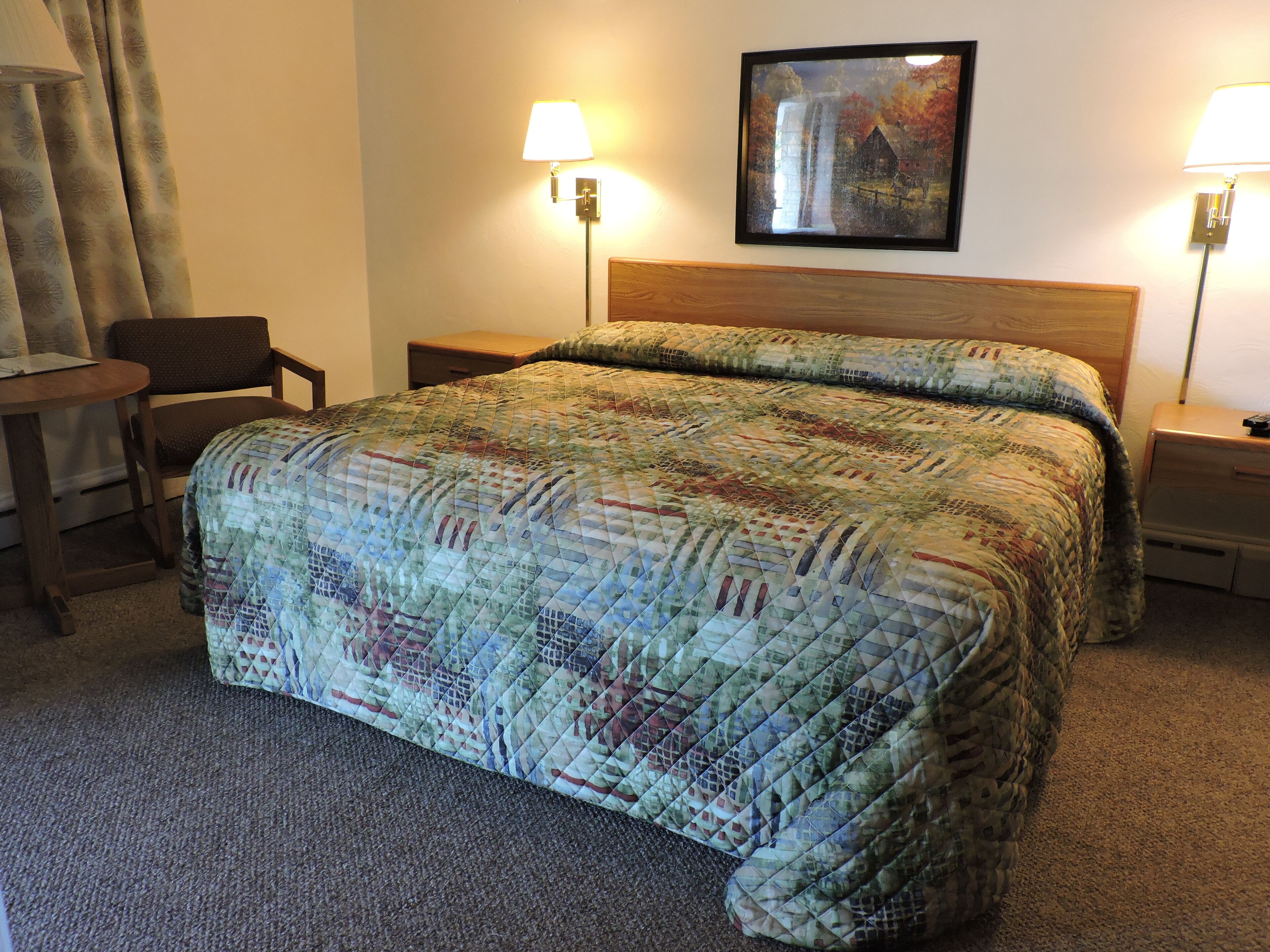 Una camera d'albergo con un letto grande, due abat-jour, una sedia, un tavolino e un quadro appeso al muro.